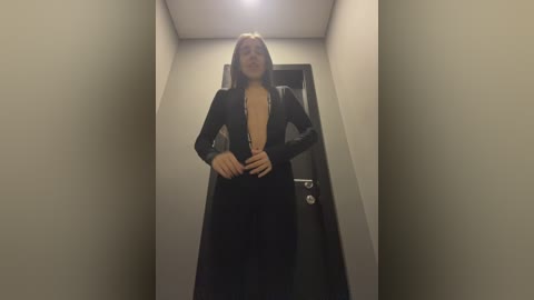 Media: Video of a woman standing in a narrow hallway. She has shoulder-length hair, fair skin, and is wearing a black, long-sleeved, open-front dress. The background features beige walls and a black door.