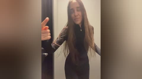 Media: Video of a young woman with long brown hair, wearing a black top and pants, giving a thumbs-up in a dimly lit, narrow hallway.