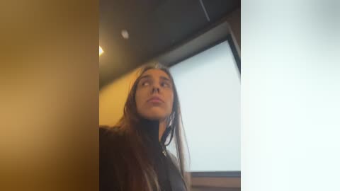 Media: A video of a young woman with long, straight brown hair and fair skin, wearing a black jacket, standing in a dimly lit room with beige walls and a large window with closed blinds.