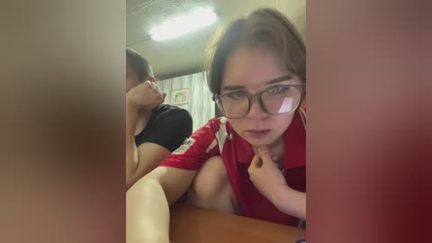 Video of a woman with glasses, wearing a red shirt, with a man in the background, blurry, in a dimly lit room with a ceiling light.