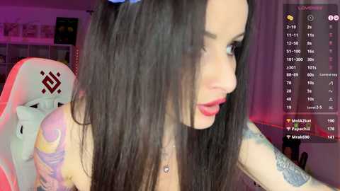 Media: Video of a pale-skinned woman with long, dark hair, wearing a blue headband, seated in a pink gaming chair. She has a colorful tattoo on her right arm and is looking to the right. The background shows a digital clock and a dark room with a TV.