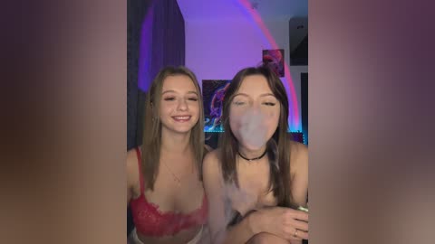 Media: Video of two young women, both with fair skin and long hair, smiling while blowing bubbles. They are wearing red lace bras, in a dimly lit room with purple lighting.