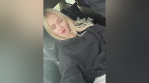 Media: Video of a blonde woman, eyes closed, sleeping in a car seat, wearing a black sweatshirt, and light-colored pants, with blurred background.