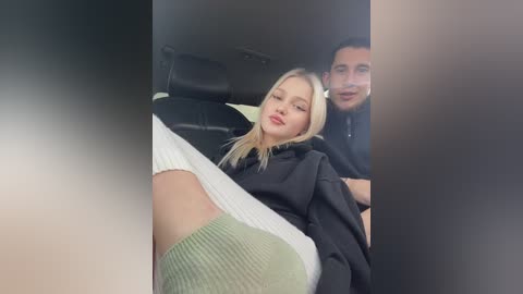 Media: Video of a blonde woman with fair skin in a black sweater, seated next to a dark-haired man in a black shirt in a car, legs visible.