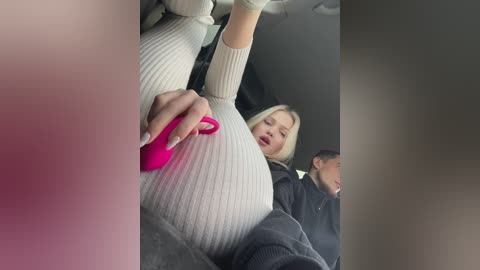 Media: A video shows a blonde woman in a white ribbed sweater and gray pants, holding a bright pink vibrator in a car. A man in a black jacket sits behind her.