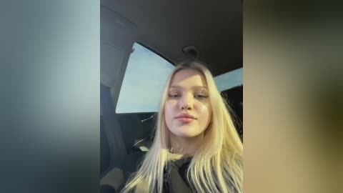 Media: A video of a young Caucasian woman with long blonde hair, fair skin, and light makeup, wearing a black jacket, sitting in a car. The background shows the car's interior.