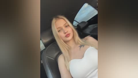 Media: Video of a blonde woman with fair skin, wearing a white strapless top, reclining in a black leather car seat, with a blue light visible in the background.