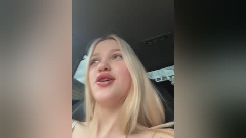 Media: Video of a blonde woman with fair skin, wearing a light-colored top, sitting in a car. Her hair is straight, and she has a neutral expression. The car's interior is visible in the background.