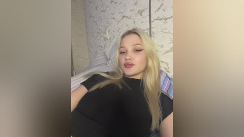 Media: Video of a young blonde woman with fair skin, wearing a black t-shirt, leaning against a textured white wall with a striped shirt draped behind her.