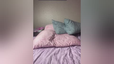 Video of a neatly made bed with light lavender sheets, two large teal pillows, and a pink pillow. The background features a textured beige wall with a small pink bouquet on the nightstand.