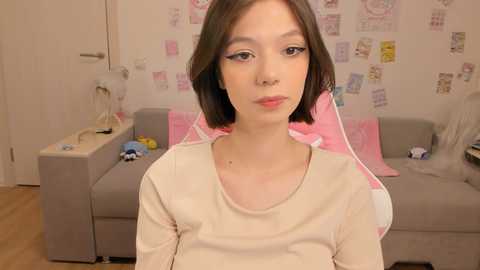 Video of a young woman with pale skin and short brown hair, wearing a beige top, sitting in a pink and white bedroom with a gray couch.