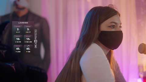 Video of a young woman with long brown hair, wearing a black face mask, white top, and black jacket, standing against a colorful, blurred background.