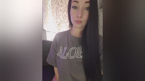 A video of a young woman with long, straight black hair, wearing a grey \"ALOHA\" t-shirt, standing in a dimly lit room with a floral-patterned curtain in the background.