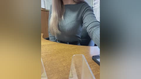 Video of a woman in a gray ribbed sweater, seated at a wooden table with a clear glass in front. She has long blonde hair and is partially obscured by the frame. Background includes a window with blinds and a wooden cabinet.