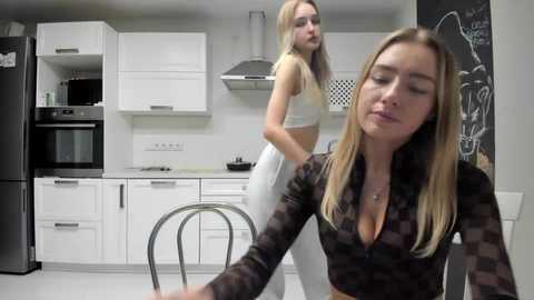 Media: Video of two young Caucasian women in a modern kitchen. One in a black crop top, the other in a white crop top and leggings.