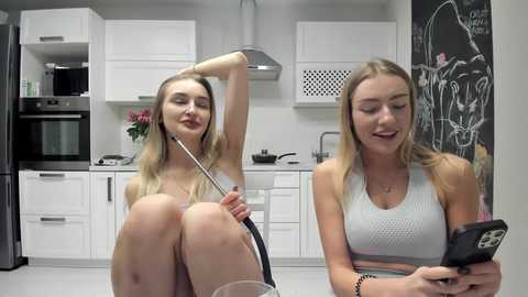 Media: Video of a blonde Caucasian woman in a white sports bra, holding a stethoscope, sitting on a kitchen counter. Another woman, also blonde, wearing a light blue tank top, is sitting next to her, smiling at her phone.