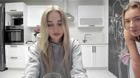 Media: Video of a young blonde woman in a gray hoodie sitting at a kitchen counter with white cabinets and black appliances. Background shows a chalkboard with drawings and a pink flower arrangement.