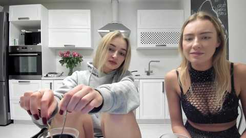 Media: Video of two young women with fair skin and blonde hair, one in a gray hoodie, the other in a black lace bra, sitting in a modern white kitchen with white cabinets and black appliances.