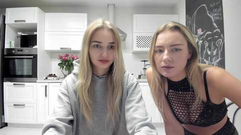 Media: Video of two young women with long blonde hair, one in a gray hoodie, the other in a black mesh top, in a modern kitchen with white cabinets and a blackboard wall.