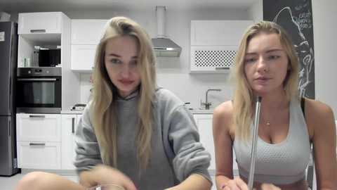 Media: Video of two blonde women in a modern kitchen; one wears a grey hoodie, the other a white tank top, both smiling.