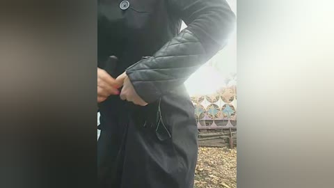 Video of a person in a black coat, adjusting the coat's collar in a dimly lit, outdoor setting with a stone wall and decorative tiles in the background.