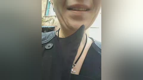 Video of a woman with a black beak prosthetic nose, smiling, wearing a black shirt with a black button, and a black choker necklace. Background shows a blurred brick wall.