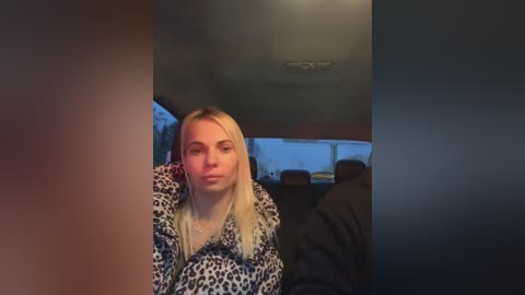 Media: Video of a young woman with long blonde hair, wearing a leopard print jacket, seated in a dimly lit, modern living room with a large window and minimalistic decor.