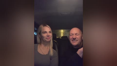 Media: Video of a smiling blonde woman in a grey top and a bald, grinning man in a black shirt seated in a dimly lit car.