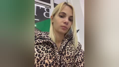 Media: Video of a young Caucasian woman with straight, platinum blonde hair, wearing a leopard print jacket, indoors near a green and white sign, looking slightly off-center.