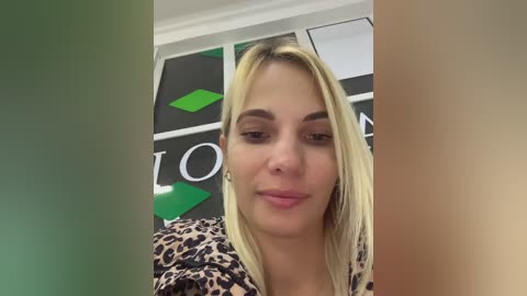 Media: Video of a blonde woman with straight hair, wearing a leopard print top, smiling softly, in a room with green and black geometric-patterned walls.