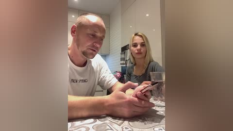 Media: Video of a bald, bearded man in a white t-shirt, sitting at a table with a blonde woman in a gray top, both holding smartphones, in a modern, well-lit kitchen with white tiles and glass cabinets.