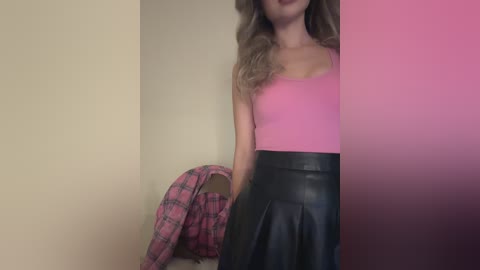 Media: A video of a fair-skinned woman with wavy blonde hair, wearing a pink tank top and a black leather skirt, standing against a beige wall, with a plaid shirt draped over her shoulder.