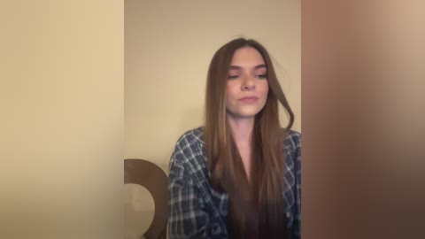 Media: Video of a young Caucasian woman with long brown hair, wearing a plaid shirt, standing against beige and brown walls, looking slightly to the side with a neutral expression.