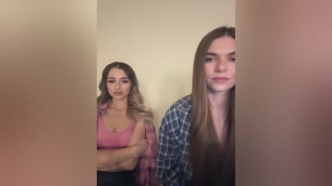 Media: Video of two young women: a blonde with long hair, wearing a pink tank top and plaid shirt, stands beside a brunette with straight hair, dressed in a black top, both facing a beige wall.