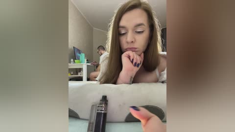 Media: Video of a young woman with light skin and long brown hair, leaning on a table, holding a black vape pen, in a casual, dimly-lit room with beige walls.