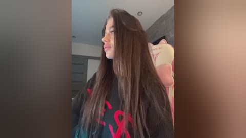 Video of a young woman with long, straight brown hair, wearing a black T-shirt with pink writing, sitting in a pink gaming chair.