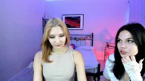 Video of two women in a bedroom: blonde in beige top, brunette in white sweater, both with neutral expressions. Background features a bed with colorful pillows, framed art, and purple and red lighting.