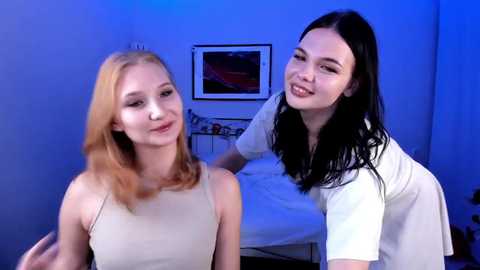Video of two young women with fair skin, blonde and brunette, smiling in a bedroom under blue light, wearing casual clothes, bed and framed artwork visible in background.