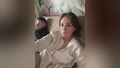 Media: A video shows a woman with medium-length brown hair and a man with short, dark hair, both in white shirts, sitting on a beige couch. The background features a large pink rose painting.
