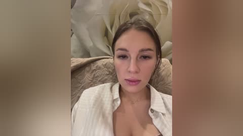 Media: Video of a young Caucasian woman with light skin and brown hair, wearing a white button-up shirt, lying on a textured beige blanket in a soft, warm-toned room.