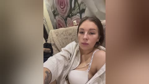 Media: A video of a young woman with light skin, brown hair, and tattoos, wearing a white bra and unbuttoned shirt, sitting on a bed with floral-patterned sheets and a beige blanket.
