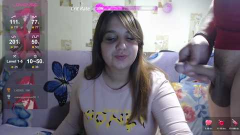 Video of a young woman with long brown hair, wearing a yellow butterfly-patterned top, sitting on a floral-patterned couch in a cozy, colorful room. A man's arm is seen on the right.