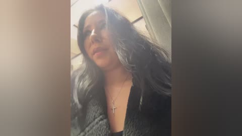 Video of a young woman with long, dark hair, wearing a black, textured cardigan, standing indoors. The background shows a beige wall and part of a ceiling. She has a neutral expression.