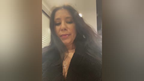 Video of a middle-aged woman with long, dark hair, wearing a black lace top and a gold necklace, smiling softly in a blurred indoor setting.