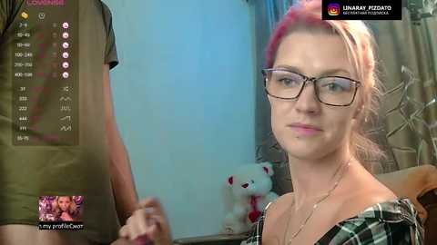 Video of a fair-skinned woman with pink hair, wearing glasses and a plaid shirt, smiling next to a man in a green shirt. Background features a plush bear and curtains.