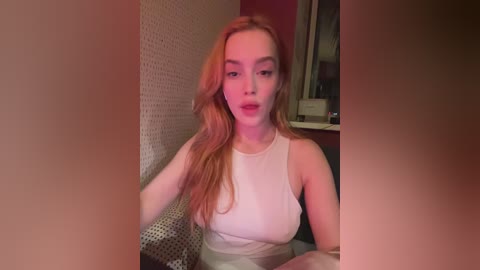 Video of a young woman with fair skin and long, wavy red hair, wearing a white tank top, sitting on a textured white wall. Background includes a mirror and a blurred room with red walls.