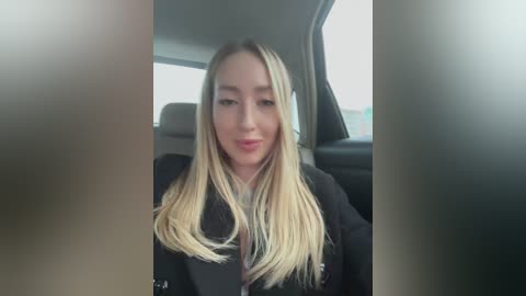 Media: A video of a young Caucasian woman with long, straight blonde hair, wearing a black jacket, sitting in a car, smiling. The background shows a blurred interior.