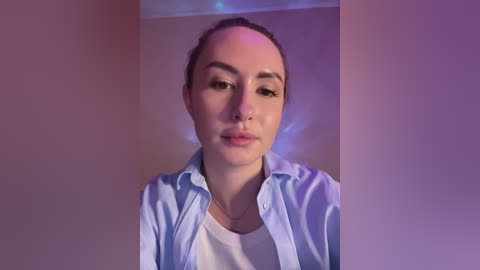 Video of a young Caucasian woman with fair skin and straight, dark brown hair tied back. She wears a light blue shirt over a white top, and has a calm, neutral expression. Background is a plain, beige wall with subtle purple lighting.