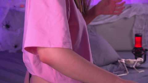 A video of a person in a pink t-shirt, possibly female, with light skin tone, holding a smartphone in a dimly lit bedroom with a bed, nightstand, and a pink lamp.