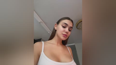 Media: Video of a young woman with light skin, brown hair, and wearing a white tank top, standing in a modern, minimalist room with white walls, a ceiling fan, and recessed lighting.